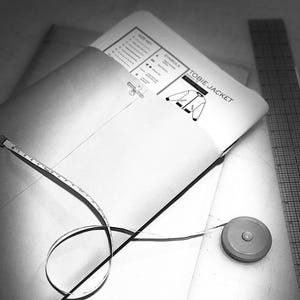 May include: A black and white image of sewing patterns and tools. The pattern envelope is open, revealing the text "TOBIE JACKET" and diagrams. A measuring tape and a ruler are also visible, suggesting a focus on garment construction and design.