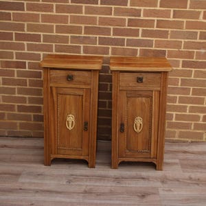 May include: A pair of wooden bedside cabinets in a natural finish. Each cabinet has a small drawer, a door with decorative hardware, and an ornate design. The cabinets are set against a brick wall.