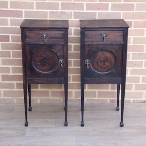 May include: A pair of antique dark wood bedside cabinets. Each cabinet features a small drawer, a circular door panel, and a decorative handle. The cabinets stand on four slender legs. The background is a brick wall.