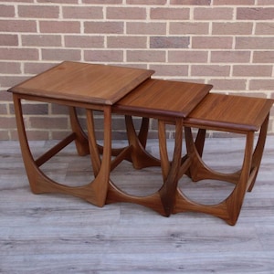 May include: A set of three nesting tables crafted from medium-toned wood. Each table features a square top and a distinctive base design with curved, arching legs that connect at the bottom, creating a continuous, open frame structure.