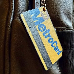 以下が含まれることがあります： 黒いストライプと青い文字でMetroCardと印刷された、黄色と青のメトロカードを模した長方形のキーホルダータグ。タグは金属製のリングに取り付けられており、ダークブラウンの質感のあるレザーの表面にぶら下がっています。