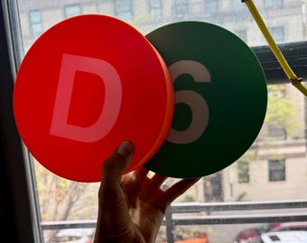May include: Two round, colorful signs are held up in front of a window. One is red with a white "D", the other is green with a white "6". The signs are circular and appear to be made of a solid material.