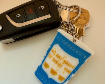 May include: A black car key fob and two brass keys attached to a white and blue coffee cup keychain. The keychain features a blue Greek key pattern at the top and the yellow text WE ARE HAPPY TO SERVE YOU with three small yellow coffee cup icons.