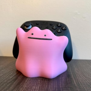 May include: A pink Ditto figure with a simple black face is in front of a black Nintendo Switch controller. The Ditto figure is smooth and rounded, with a playful appearance. The controller has multiple buttons and joysticks.