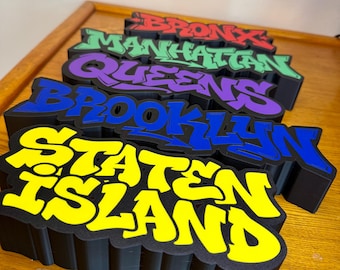 May include: A set of five stacked 3D signs with black bases and colorful graffiti-style lettering. The signs display the names of the five New York City boroughs: Bronx in red, Manhattan in green, Queens in purple, Brooklyn in blue, and Staten Island in yellow.