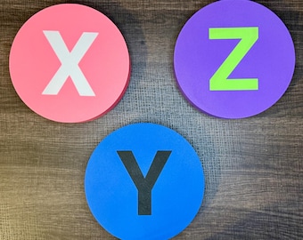 May include: Three circular foam discs featuring letters. A pink disc displays a white letter X, a purple disc features a lime green letter Z, and a blue disc shows a black letter Y. Each disc has a smooth, flat surface and a uniform, rounded shape.