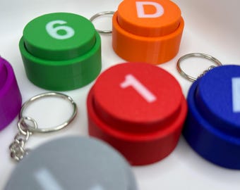 May include: A collection of colorful circular keychains, each featuring a raised button-like top with a white character printed on the surface. The set includes green with the number 6, orange with the letter D, red with the number 1, blue with the letter A, purple, and gray.