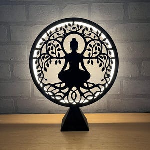 Op de afbeelding: Een zwarte, cirkelvormige decoratieve lamp met een silhouet van een zittende Boeddha-figuur in een levensboomontwerp. De lamp heeft een helder, wit licht dat van achter het ontwerp komt en staat op een zwarte, driehoekige basis. De achtergrond is een grijze bakstenen muur.