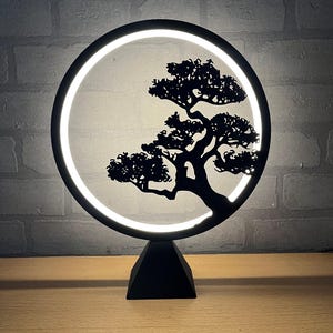 Op de afbeelding: Een zwarte LED-lamp met een silhouet van een bonsai-boom in een cirkelvormig frame. De boom is gedetailleerd en het frame straalt zacht, wit licht uit. De lamp staat op een zwarte, geometrische basis. Geschatte hoogte: 25 cm.