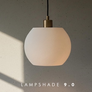 Op de afbeelding: Een bolvormige witte lampenkap met een textuur van diagonale ribbels, opgehangen aan een zwart snoer aan een messingkleurige metalen armatuur. De tekst LAMPSHADE 9.0 staat in het wit onderaan in het midden van de afbeelding.