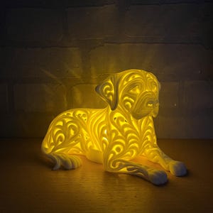 May include: A glowing, cream-colored dog-shaped lamp with intricate, swirling cut-out patterns. The lamp is in a resting position, with its paws extended. The light emanates from within, creating a warm, inviting ambiance. The lamp is on a wooden surface.