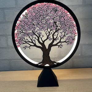 Op de afbeelding: Een zwart, cirkelvormig, verlicht kunstwerk van een kersenbloesemboom. De stam en takken van de boom zijn bruin, met roze en witte bloesems. De boom staat tegen een heldere achtergrond in een zwart cirkelvormig frame, gemonteerd op een zwarte piramidebasis.