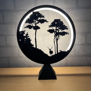Op de afbeelding: Een zwarte, ronde decoratieve lamp met een silhouet van een boslandschap met bomen en een hert. De lamp heeft een helder, wit licht in het midden en staat op een zwarte, geometrische basis. Het ontwerp is minimalistisch en geïnspireerd op de natuur.