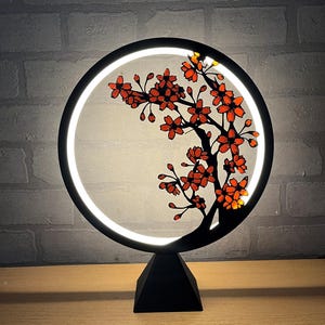 Op de afbeelding: Een zwarte, cirkelvormige LED-lamp met een kersenbloesem ontwerp. De lamp heeft een witte lichtring en een zwarte basis. Het kersenbloesem ontwerp is in tinten rood en oranje.