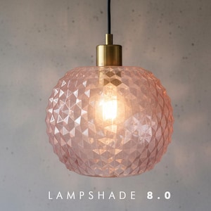 Pode incluir: Uma luminária pendente esférica de vidro rosa claro com uma textura geométrica facetada. A luminária possui um suporte de metal dourado no topo, um cabo preto e uma lâmpada iluminada no interior. O texto "LAMPSHADE 8.0" está impresso na parte inferior.