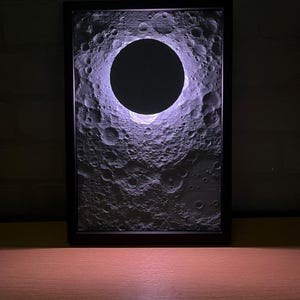 Peut inclure: Une œuvre d'art encadrée et éclairée représentant un relief détaillé de la surface lunaire, avec un grand vide circulaire sombre au centre. L'œuvre est encadrée dans un cadre noir, et la surface lunaire est texturée avec des cratères.