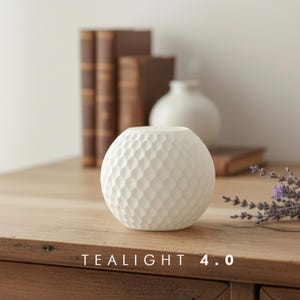 Peut inclure: Un bougeoir chauffe-plat sphérique en céramique blanche avec un motif en nid d'abeille texturé sur sa surface extérieure. La base de l'objet est plate et le sommet présente une ouverture circulaire. Le texte "TEALIGHT 4.0" est imprimé en blanc en bas.
