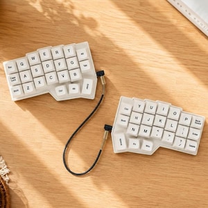Peut inclure: Un clavier ergonomique séparé avec des touches blanches et des lettres noires. Les deux moitiés sont reliées par un câble noir en spirale avec des connecteurs métalliques. La disposition comprend des touches QWERTY, de modification et de fonction.