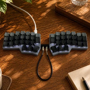 Pode incluir: Um teclado mecânico ergonómico dividido em duas secções separadas de teclas pretas com retroiluminação branca. As duas metades estão ligadas por um cabo preto em espiral e um cabo de alimentação branco sai da parte superior esquerda.