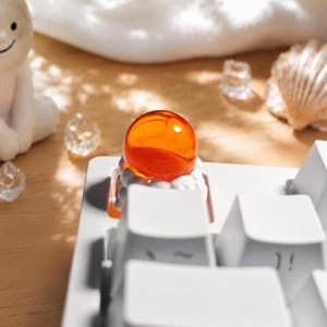 Può includere: Un tasto personalizzato per tastiera meccanica con una sfera arancione traslucida poggiata su una base a forma di nuvola bianca scolpita. Il tasto è installato su una tastiera bianca, circondato da piccoli cristalli decorativi trasparenti e una conchiglia.
