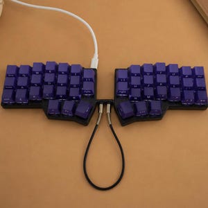 May include: A split ergonomic mechanical keyboard featuring dark purple keycaps with white lettering. The two halves are connected by a black coiled cable, and a white USB cable extends from the top of the left unit. The base is black.
