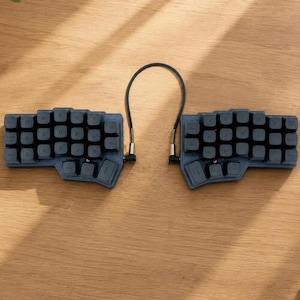 May include: A dark grey split mechanical keyboard consisting of two separate keypads connected by a black cable. Each keypad features a contoured, ergonomic layout with dark grey keys displaying white letters and symbols in a QWERTY arrangement.