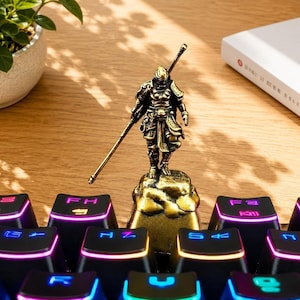 Könnte beinhalten: Eine goldfarbene Metallfigur eines Kriegers in detaillierter Rüstung, der einen langen Stab hält und auf einem felsförmigen Sockel steht. Die Figur ist als individuelle Tastenkappe auf einer schwarzen mechanischen Tastatur mit RGB-Beleuchtung.