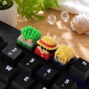 Può includere: Tre tasti personalizzati per tastiera meccanica con teste di personaggi in stile anime scolpite. Uno ha capelli verdi a punta, uno indossa un cappello di paglia giallo con una fascia rossa e uno ha capelli biondi con un dettaglio a spirale.
