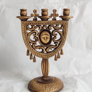 May include: A carved wooden menorah with a detailed design. The menorah has seven candle holders, a central decorative panel with a face, and a round base. The wood has a warm, brown tone.