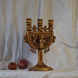 May include: A carved wooden candelabra featuring three candle holders, each topped with a sculpted human face. The central structure displays an intricate openwork design with a central figure, decorative floral motifs, and small hanging tassels.