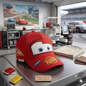 Può includere: Un cappellino da baseball rosso brillante che raffigura il personaggio Saetta McQueen del film Cars. Il cappellino presenta grandi occhi bianchi sul davanti, un fulmine giallo sul lato e un logo Rust-eze sulla visiera. Un'etichetta riporta la scritta Handmade for Little Racers.