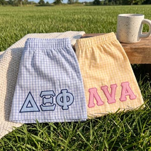 May include: Two pairs of gingham shorts with elastic waistbands. The left pair is light blue and white with dark blue Greek letters embroidered on the front. The right pair is yellow and white with pink Greek letters embroidered on the front.