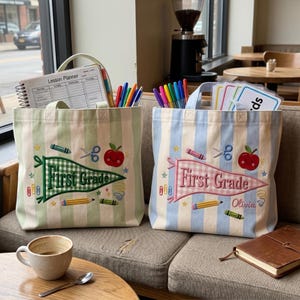May include: Two striped canvas tote bags featuring embroidered school-themed designs. One bag has a green gingham pennant with the text First Grade, while the other has a pink gingham pennant with First Grade and the name Olivia embroidered.