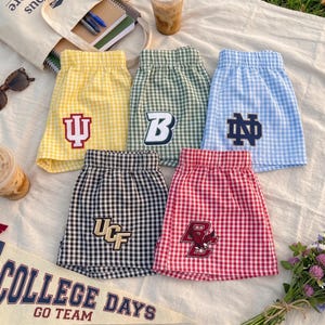 May include: Five pairs of gingham shorts in yellow, green, light blue, black, and red. Each pair features a unique embroidered collegiate logo on the front, including the IU, B, ND, UCF, and BC eagle emblems, with elasticized waistbands.