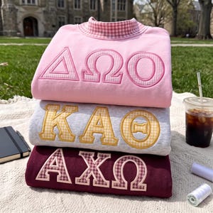 May include: A stack of three crewneck sweatshirts featuring Greek letters appliqued in gingham patterns. The top is light pink with pink gingham letters, the middle is heather gray with yellow gingham letters, and the bottom is maroon with maroon gingham letters.