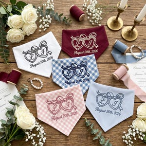 May include: Five triangular fabric bandanas featuring embroidered twin hearts with the letters L and J, and the text August 20th, 2026. The bandanas come in white, burgundy, blue gingham, pink gingham, and blue pinstripe patterns.