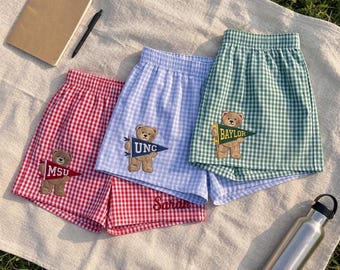 May include: Three pairs of gingham shorts in red, light blue, and green. Each pair features an embroidered teddy bear holding a pennant flag labeled MSU, UNC, or BAYLOR. The red shorts have the name Sarah embroidered in red script near the hem.