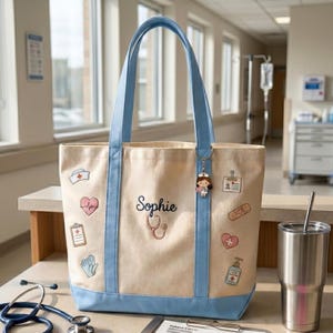 May include: A beige canvas tote bag with light blue trim and handles. The bag features the name Sophie embroidered in black, a stethoscope graphic, and various medical-themed icons including a nurse hat, heart, clipboard, gloves, and sanitizer.