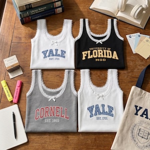 May include: Four sleeveless tank tops with lace trim and small bows at the neckline. Two white tops feature blue Yale text, one black top displays yellow University of Florida text, and one gray top shows red Cornell text with established dates.