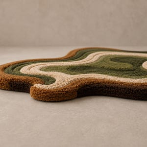 May include: An irregularly shaped tufted rug featuring a series of concentric, wavy bands in shades of dark brown, rust orange, cream, olive green, and forest green. The rug has a thick, textured pile surface with organic, flowing contours.