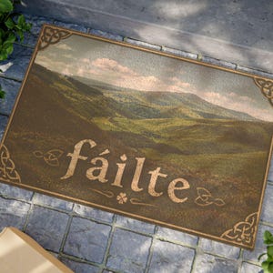 May include: Rectangular doormat featuring a landscape of green hills under a cloudy sky. The word "fáilte" is printed in a decorative font. The design includes corner and border embellishments.