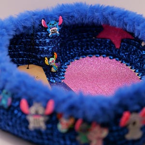 Op de afbeelding: Een blauwe gehaakte mand met een roze stervormig accent en Stitch-karakterversieringen. De mand heeft een pluizige blauwe rand en is versierd met verschillende Stitch-beeldjes, wat een grillig en kleurrijk ontwerp creëert.