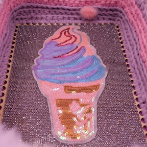 Può includere: Un cestino portaoggetti quadrato con pareti all'uncinetto rosa e viola e un bordo bianco soffice. La base presenta una superficie viola glitterata con una grande toppa centrale a forma di cono gelato decorata con paillettes blu, viola e rosa.