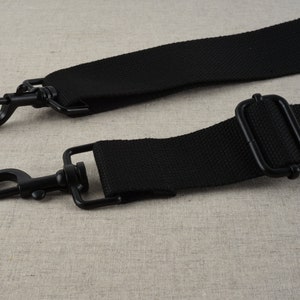 May include: Black adjustable strap with two metal clasps. The strap is made of a woven fabric.
