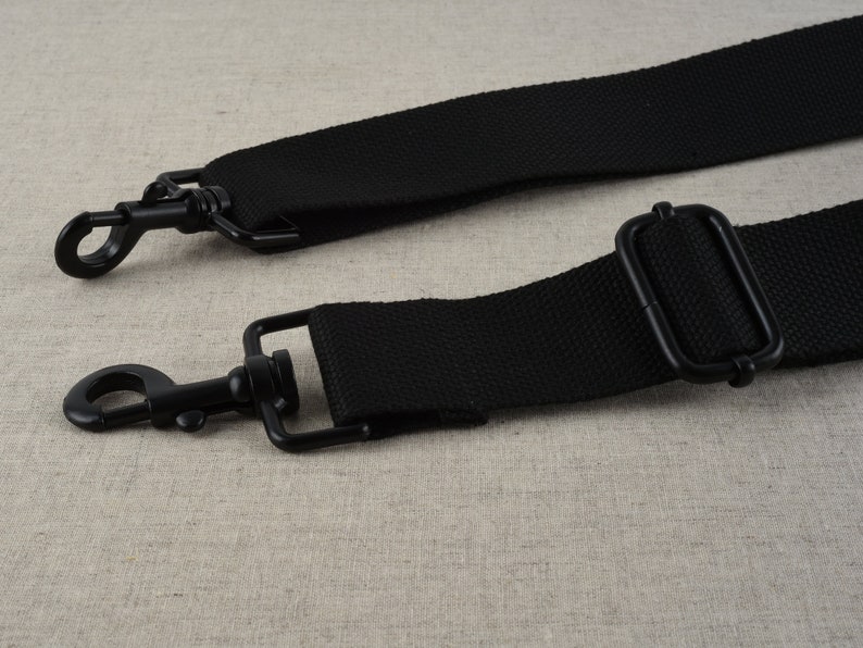 May include: Black adjustable strap with two metal clasps. The strap is made of a woven fabric.