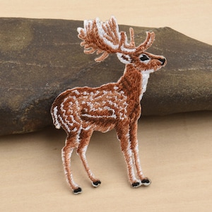 Könnte beinhalten: Gestickter Aufnäher eines Hirsches mit braun-weißem Fell, detailliertem Geweih und schwarzen Hufen. Der Hirsch steht auf einer neutralfarbenen Oberfläche. Der Aufnäher ist wahrscheinlich für Kleidung oder Accessoires.