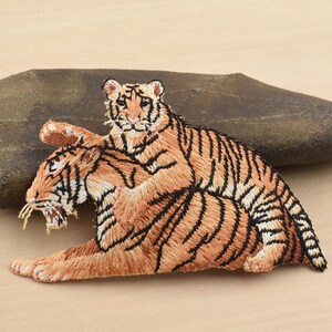 May include: Embroidered patch depicting two tigers. The tigers are orange with black stripes, and the patch is detailed with various shades of brown. One tiger is lying down, and the other is on top.