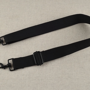 May include: Black adjustable strap with two metal clasps. The strap is made of a woven fabric.