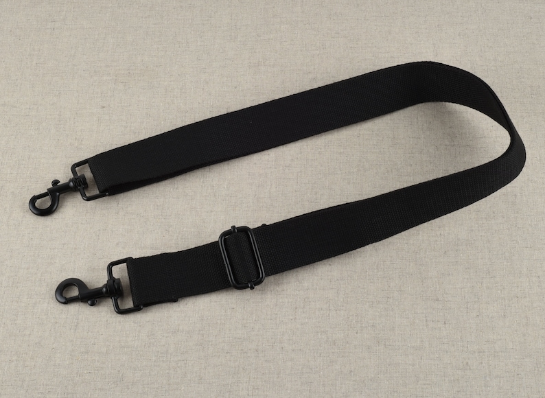May include: Black adjustable strap with two metal clasps. The strap is made of a woven fabric.