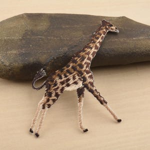 Puede incluir: Parche bordado de jirafa con un patrón de manchas marrones y beige. La jirafa tiene patas largas y una cola curvada. El parche es probablemente para ropa o accesorios.